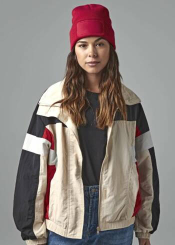 Frau in modischer Streetwear mit bunter Retro-Windjacke, schwarzem Shirt, blauer Jeans und roter Beanie-Mütze.