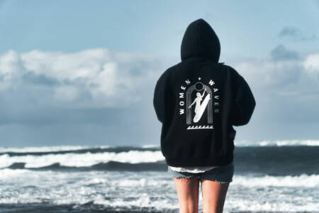 Person am Strand mit schwarzem Hoodie, darauf das Design "Women + Waves" mit Surfmotiv.