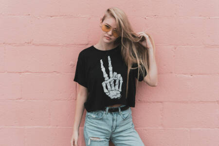 Junge Frau mit schwarzen Crop-Top und Skelett-Peace-Handmotiv posiert vor einer rosa Wand.
