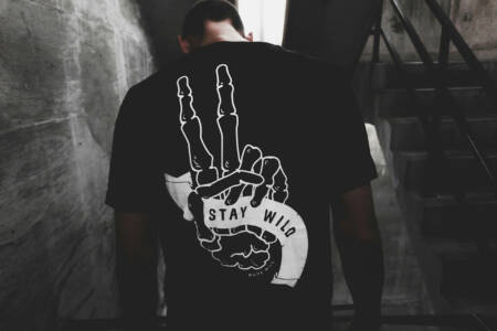 Mann in dunklem Treppenhaus mit schwarzem Shirt, auf dem ein Skelett-Handmotiv mit "Stay Wild" steht.