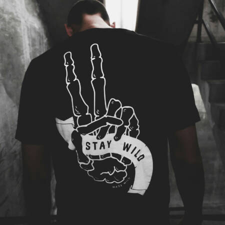 Mann in dunklem Treppenhaus mit schwarzem Shirt, auf dem ein Skelett-Handmotiv mit "Stay Wild" steht.